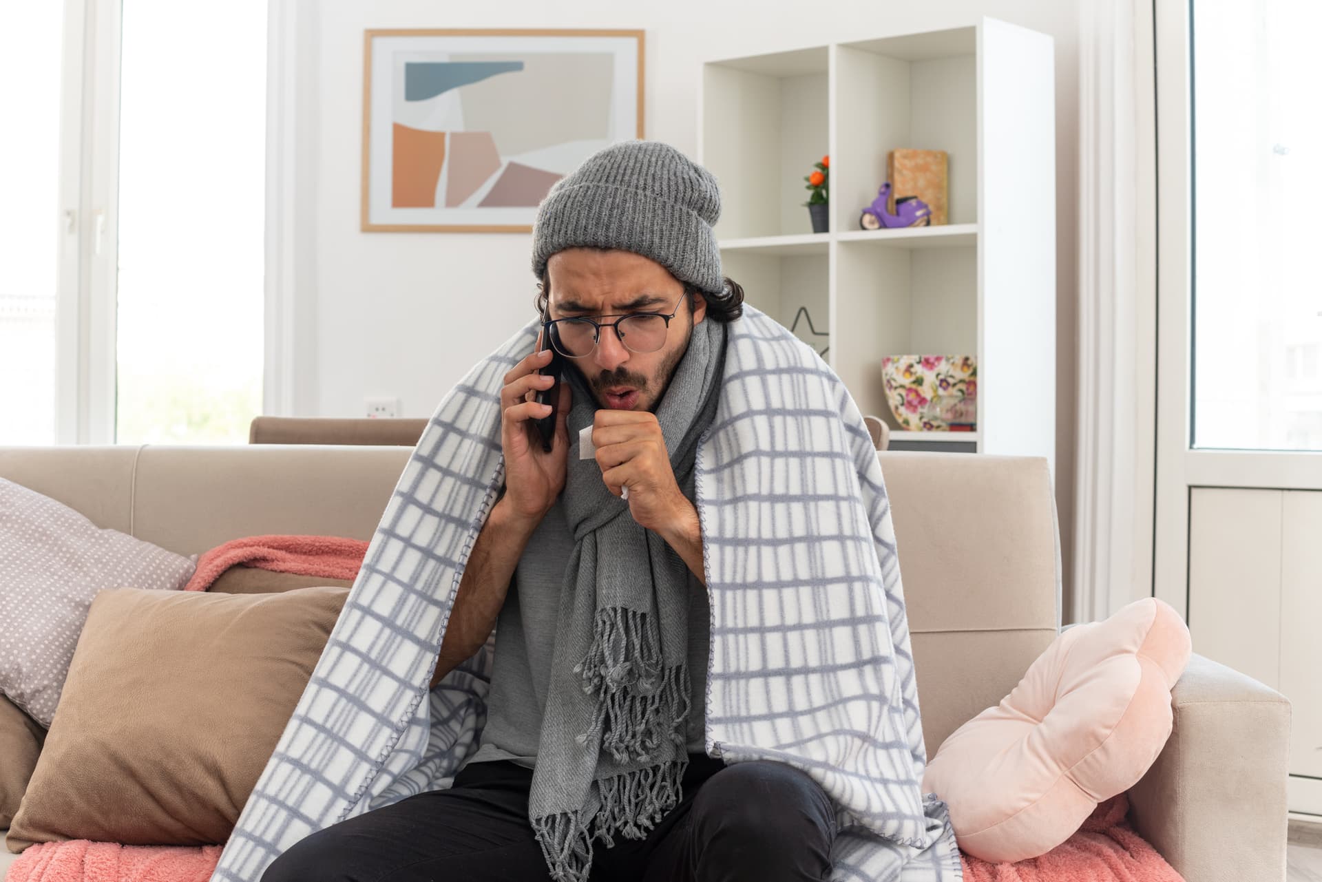 sick-young-man-optical-glasses-wrapped-plaid-with-scarf-around-his-neck-wearing-winter-hat-coughing-talking-phone-sitting-couch-living-room.jpg